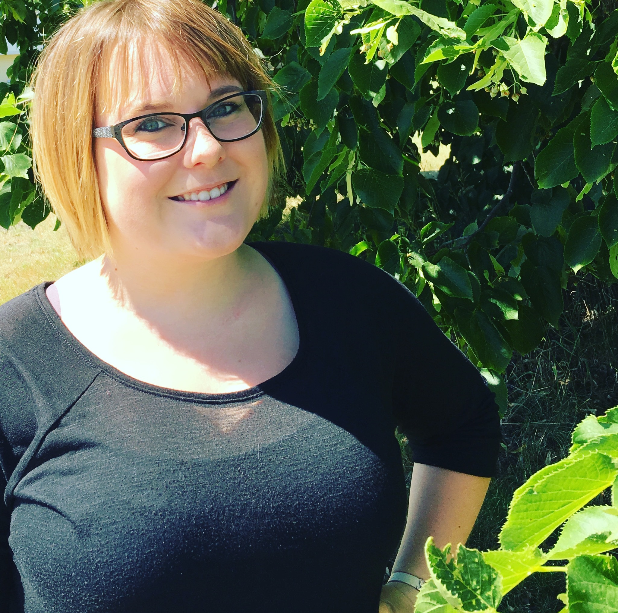 A smiling woman with glasses stands outdoors near green foliage.